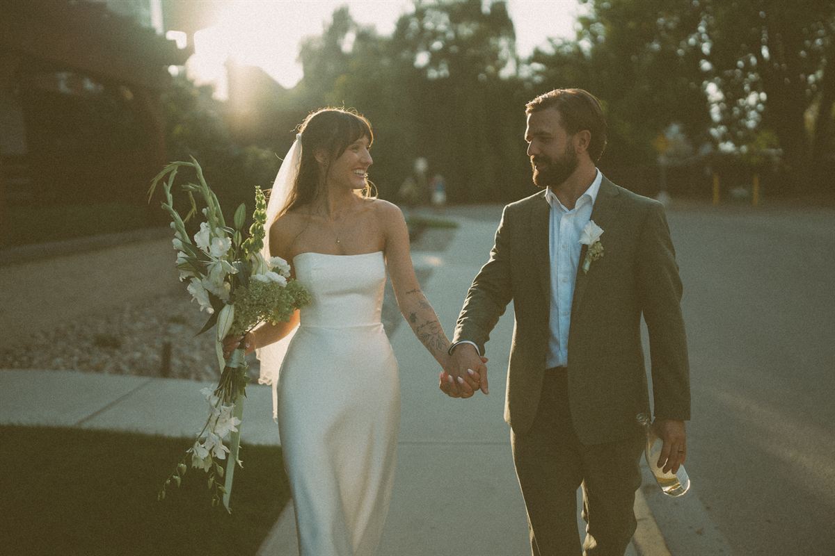 Bride and groom walking hand in hand down a Kelowna street at sunset, her bouquet in one hand and champagne in the other