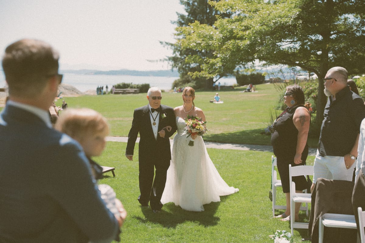 Charlotte walking the aisle with her father, the ocean at Saxe Point behind them