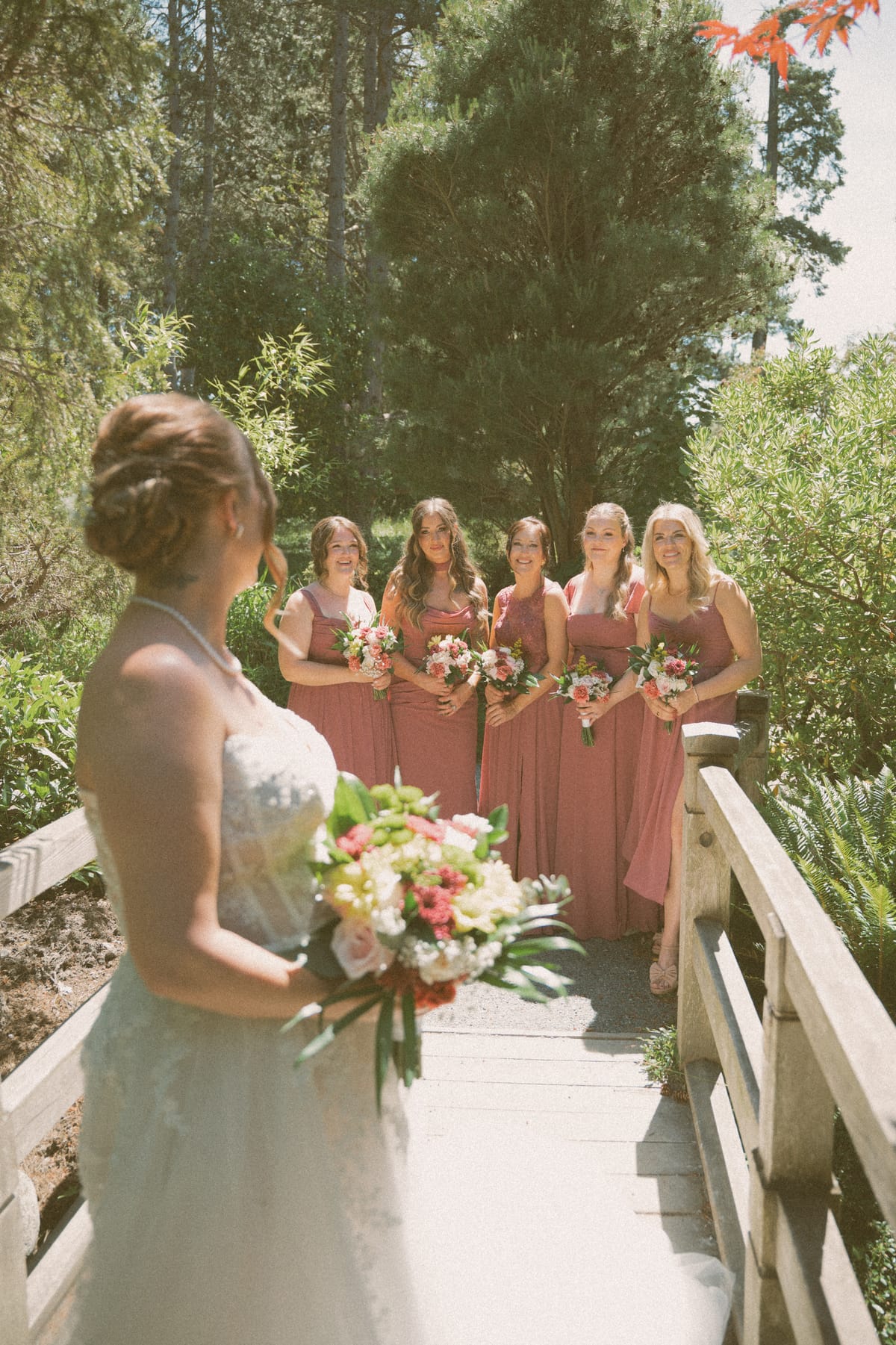 Charlotte facing her bridesmaids across a wooden footbridge in the garden