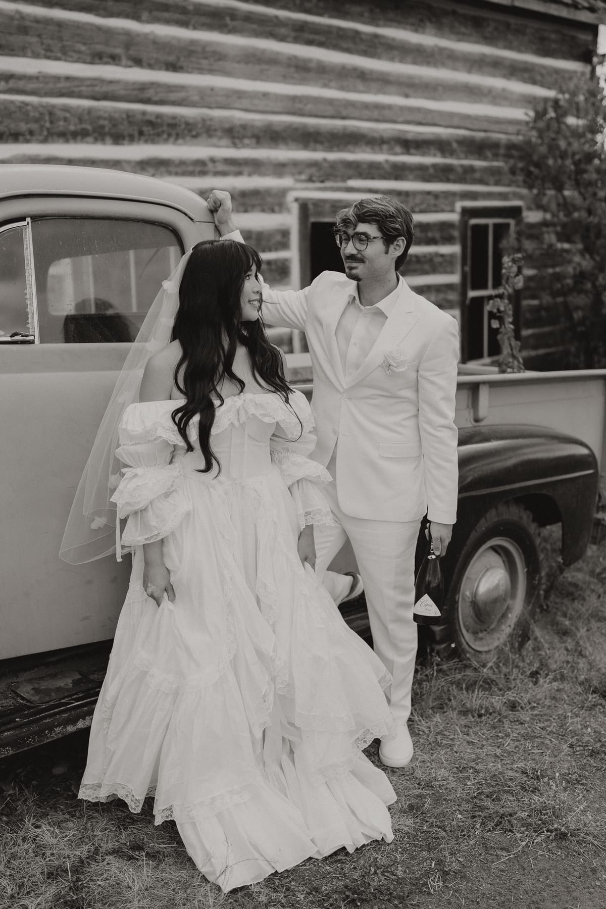 Black and white of Nina and Brandon beside a vintage truck at a log cabin