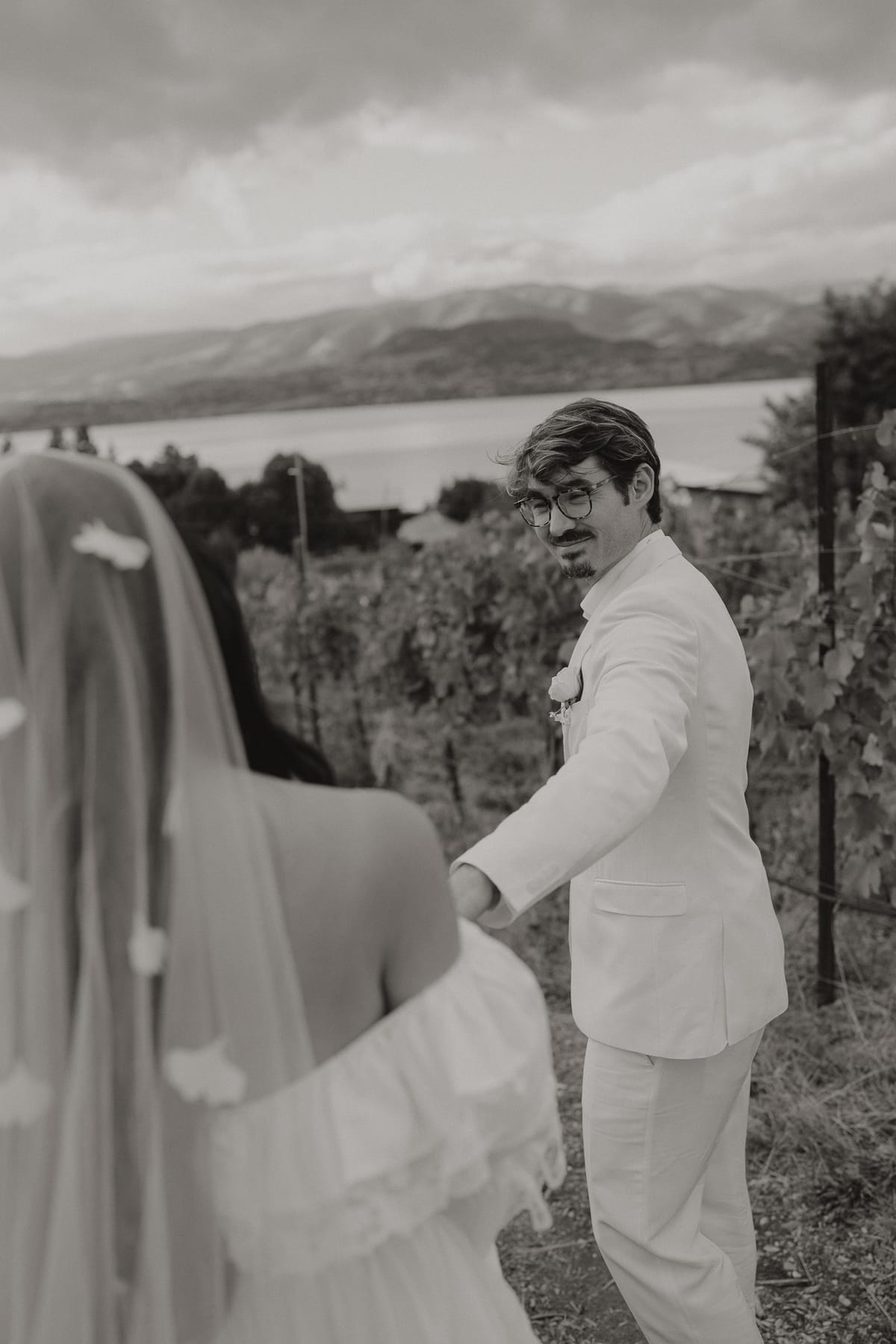 Black and white of Brandon leading Nina through the vineyard rows with the lake behind