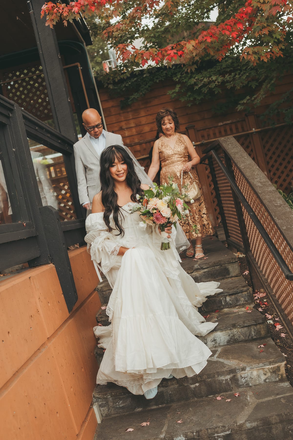Nina walking down the stone steps with her parents, bouquet in hand, autumn leaves overhead