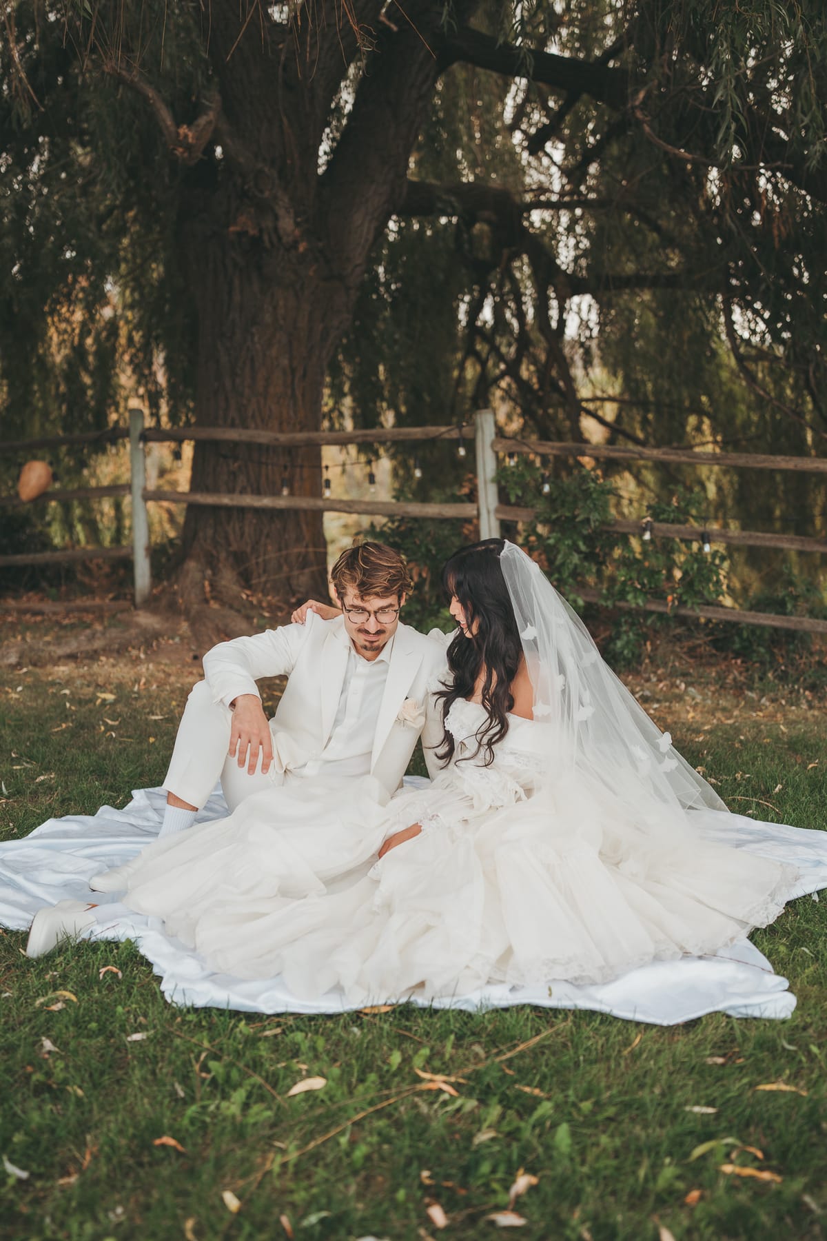 Nina and Brandon seated on a blanket beneath a willow tree, her gown spread around them