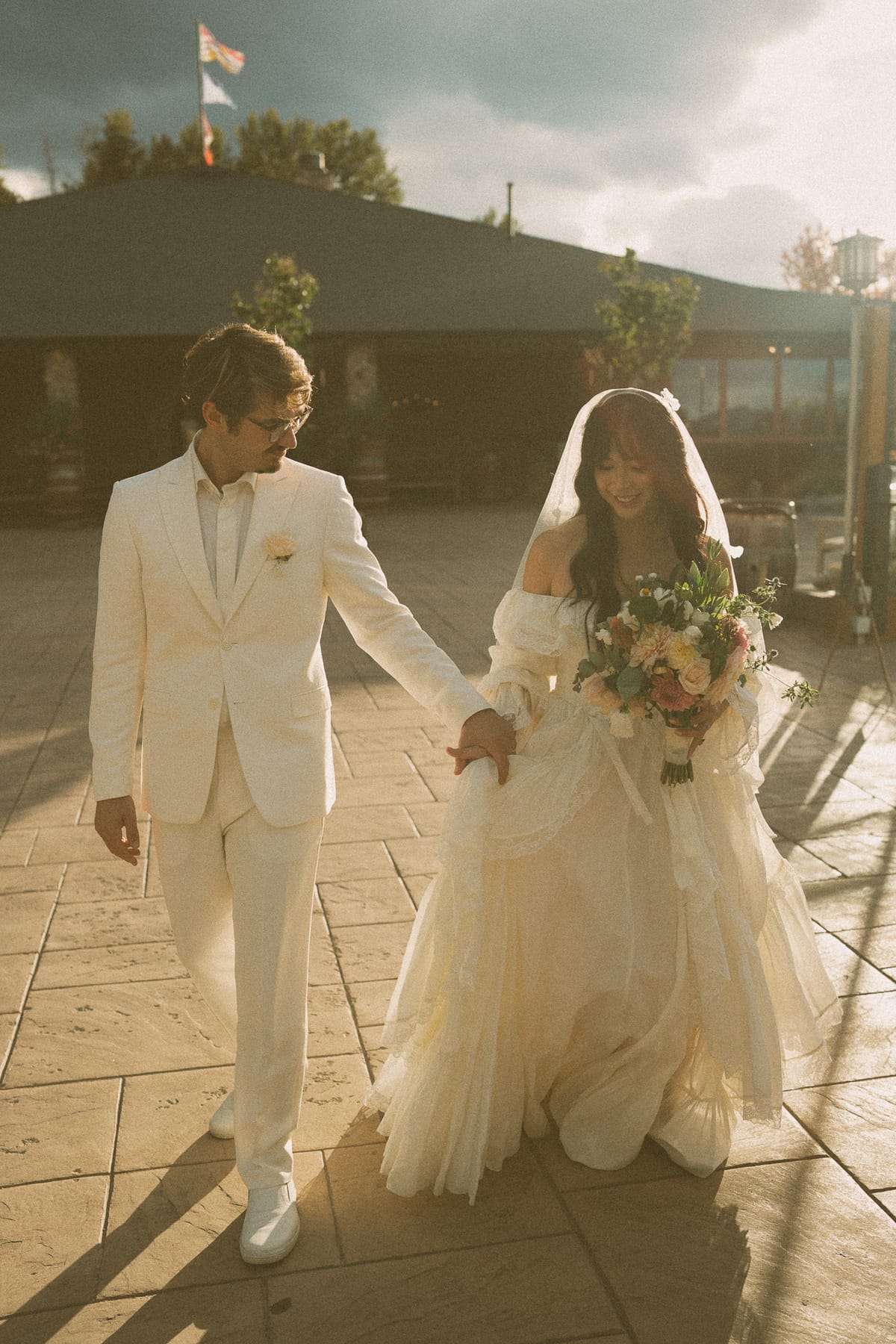 Brandon leading Nina across the sunlit courtyard at golden hour, her bouquet in hand