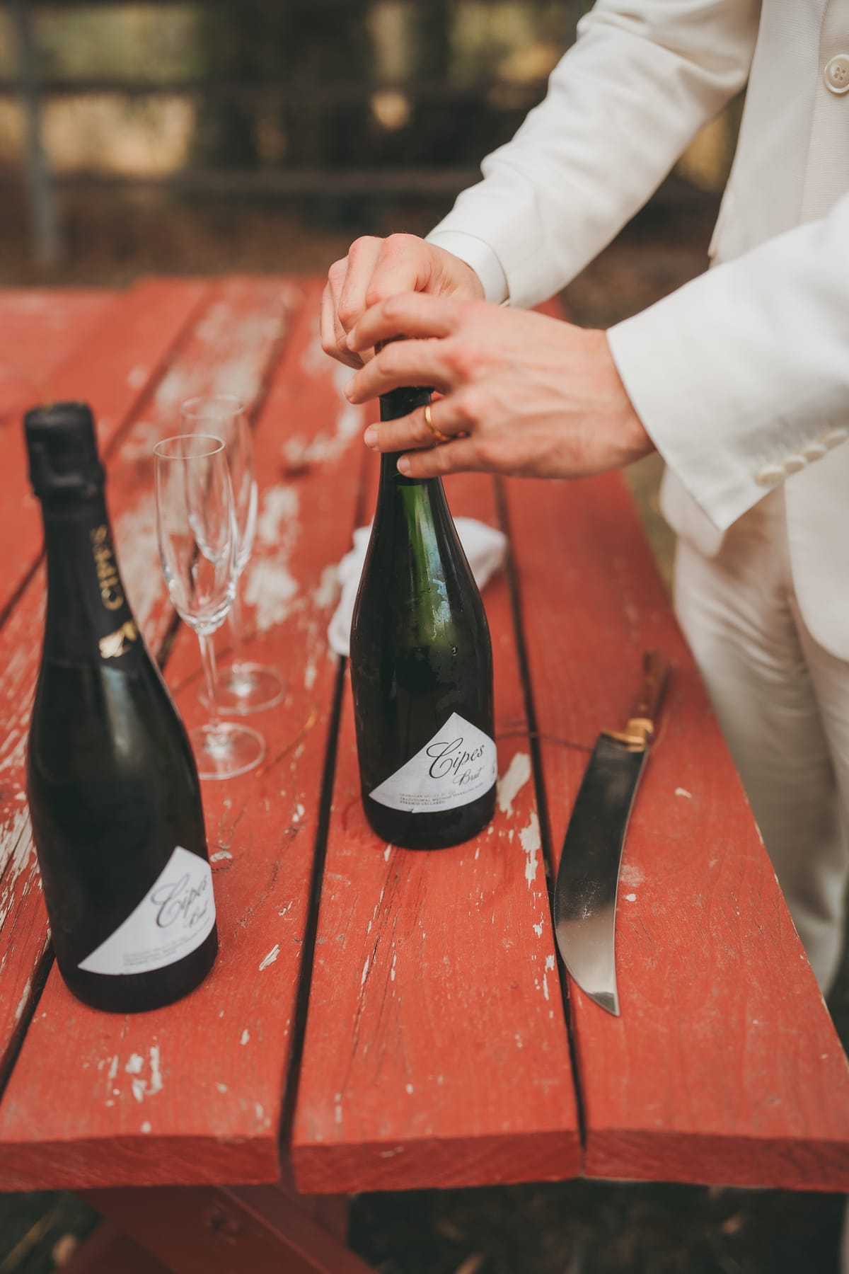 Brandon opening a bottle of sparkling wine on the red picnic table beside the flutes