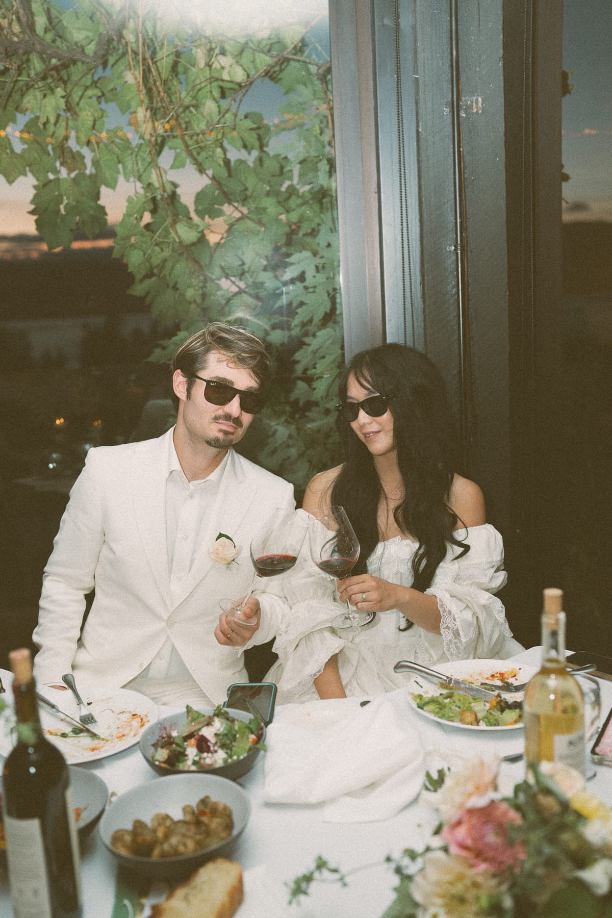 Nina and Brandon relaxing in sunglasses at their reception table with red wine