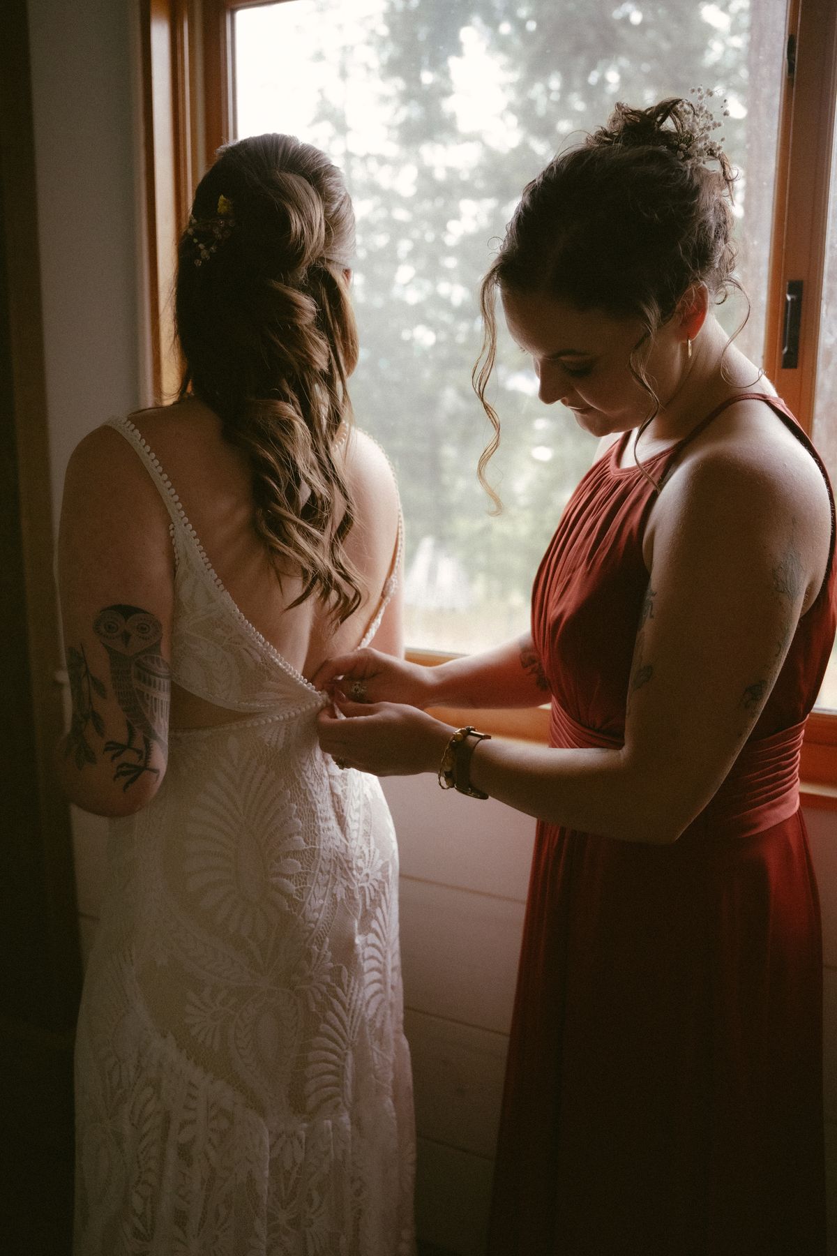 A bridesmaid in red helping the bride into her gown by the window