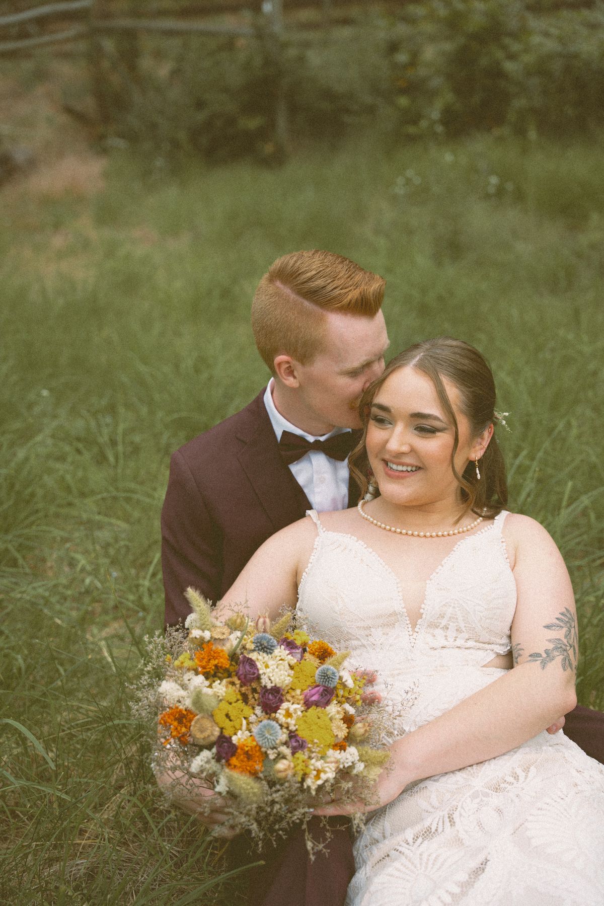 Groom kissing the bride's temple as they sit in the meadow