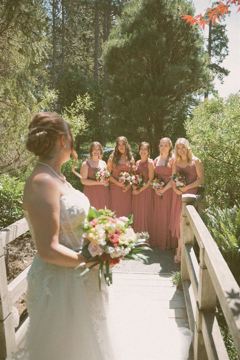 Bride watching her bridesmaids from a wooden footbridge, all of them in dusty rose beneath tall pines