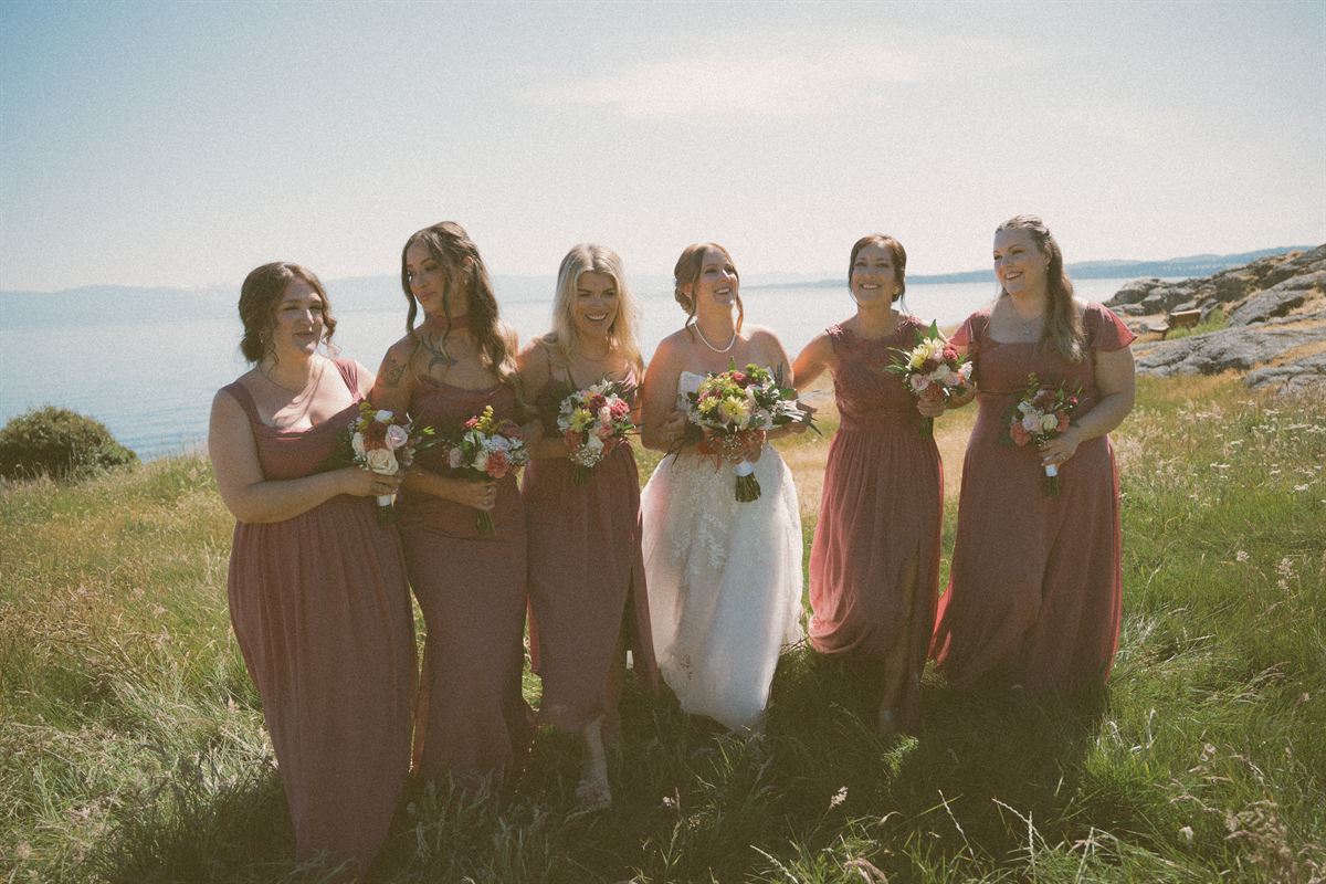 Bride and her bridesmaids laughing together in dry coastal grass on Vancouver Island, the ocean and distant islands behind them