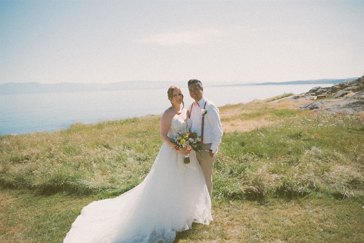 Bride and groom on a grassy coastal bluff on Vancouver Island, the ocean and far shoreline stretching out behind them